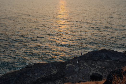 View of the sun s golden rays reflecting off the tranquil ocean waters as figures stand on the rocky shore Chiplun Maharashtra India. View of the sun s golden rays reflecting off the tranquil ocean waters as figures stand on the rocky shore Chiplun Maharashtra India.