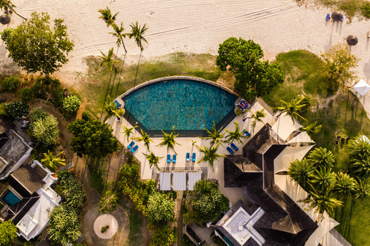 Aerial view of a luxury resort featuring a semi circular swimming pool with people swimming surrounded by palm trees and a sandy beach in Mauritius. Aerial view of a luxury resort featuring a semi circular swimming pool with people swimming surrounded by palm trees and a sandy beach in Mauritius.