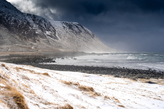 View of a snow covered coastline with dramatic dark clouds over the mountains and rocky beach in Leknes Lofoten Norway.