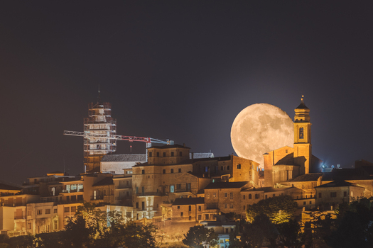 View of the town at night with a large full moon rising behind the historic buildings and church tower featuring a construction crane in Corridonia Marche Italy. View of the town at night with a large full moon rising behind the historic buildings and church tower featuring a construction crane in Corridonia Marche Italy.