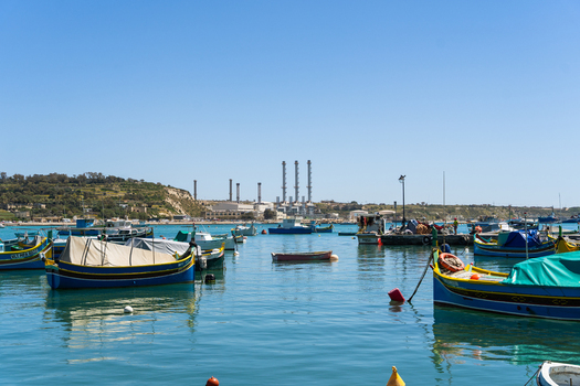 View of traditional Maltese luzzu fishing boats moored in the harbor with the Delimara Power Station chimneys in the background Marsaskala Southern Region Malta. View of traditional Maltese luzzu fishing boats moored in the harbor with the Delimara Power Station chimneys in the background Marsaskala Southern Region Malta.