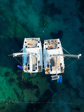 Aerial view of two catamarans moored in clear turquoise water with people relaxing on deck and paddleboards floating nearby in Split Dalmatia County Croatia.