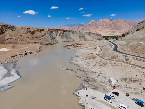 Aerial view of the Indus River winding through the arid mountain landscape of Ladakh with a road running along the bank and vehicles parked near the riverbank in Ladakh Division Ladakh India. Aerial view of the Indus River winding through the arid mountain landscape of Ladakh with a road running along the bank and vehicles parked near the riverbank in Ladakh Division Ladakh India.