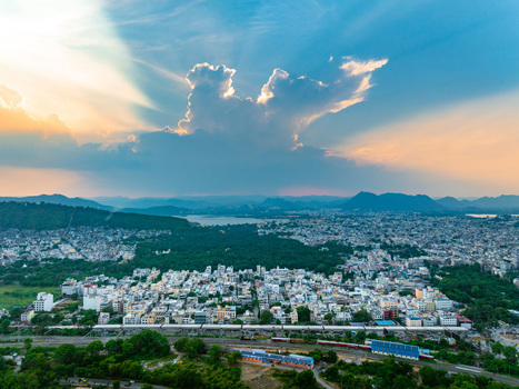 Aerial view of the cityscape with lush green hills a serene lake in the distance and a dramatic sunset sky with golden light rays over Udaipur Rajasthan India. Aerial view of the cityscape with lush green hills a serene lake in the distance and a dramatic sunset sky with golden light rays over Udaipur Rajasthan India.