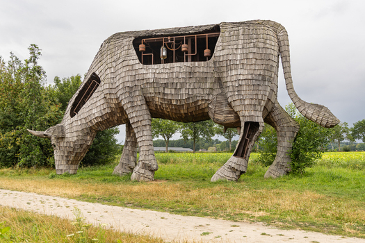 View of the large wooden cow sculpture known as De Melkfabriek featuring intricate shingle construction and open structural details in a rural field in Esbeek Noord Brabant Netherlands.