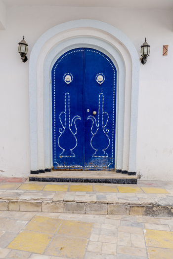 View of a traditional blue arched door with intricate white nail patterns and wall lamps set in a white wall in Kairouan Kairouan Governorate Tunisia.