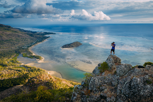 Aerial view of a photographer standing on a rocky mountain peak overlooking the turquoise lagoon and lush tropical coastline in Moka District Mauritius. Aerial view of a photographer standing on a rocky mountain peak overlooking the turquoise lagoon and lush tropical coastline in Moka District Mauritius.