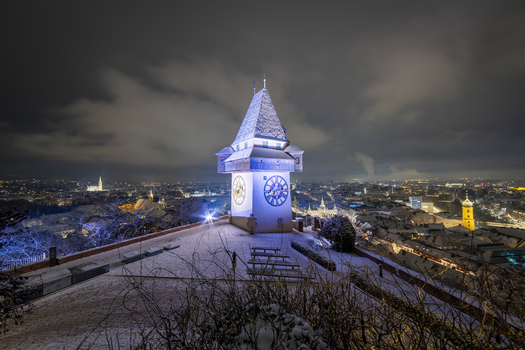 View of the illuminated clock tower on Schlossberg hill stands majestically against the night sky with the snowy cityscape twinkling below Graz Styria Austria. View of the illuminated clock tower on Schlossberg hill stands majestically against the night sky with the snowy cityscape twinkling below Graz Styria Austria.