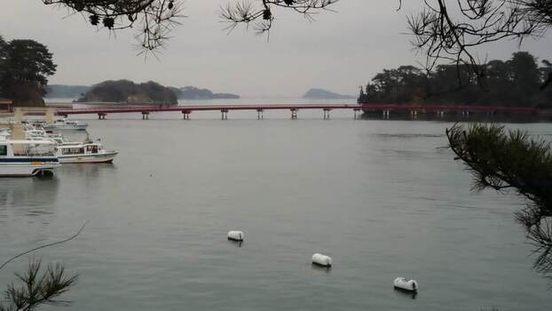 View of the red Fukuura Bridge spanning the calm waters of Matsushima Bay with small boats moored at the shoreline in Matsushima Miyagi Japan.