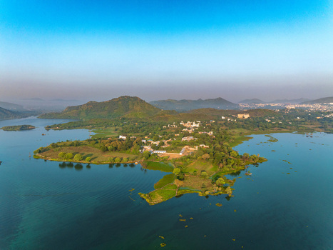 Aerial view of a lush green peninsula surrounded by the calm waters of a lake with rolling hills in the background in Udaipur Rajasthan India. Aerial view of a lush green peninsula surrounded by the calm waters of a lake with rolling hills in the background in Udaipur Rajasthan India.