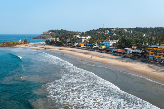 Aerial view of the sandy coastline of Kovalam Beach with turquoise waves crashing on the shore and palm trees lining the waterfront buildings under a clear blue sky Kovalam Kerala India. Aerial view of the sandy coastline of Kovalam Beach with turquoise waves crashing on the shore and palm trees lining the waterfront buildings under a clear blue sky Kovalam Kerala India.