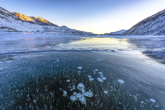 View of methane bubbles trapped in the frozen surface of a mountain lake surrounded by snow covered peaks under a clear sky in Madesimo Lombardy Italy. View of methane bubbles trapped in the frozen surface of a mountain lake surrounded by snow covered peaks under a clear sky in Madesimo Lombardy Italy.