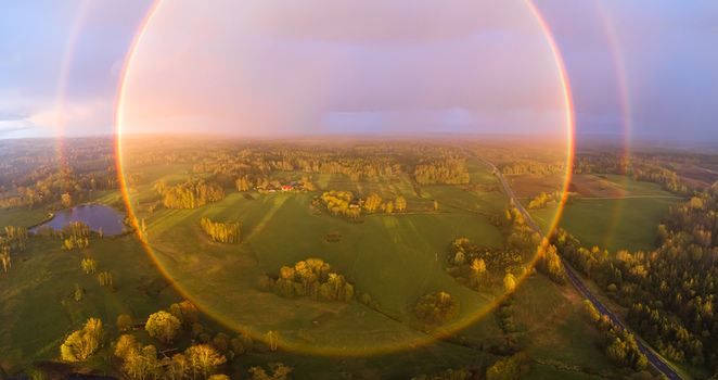 Aerial view of a full circle rainbow over the lush green fields forests and a small pond in the rural landscape of Latvia. Aerial view of a full circle rainbow over the lush green fields forests and a small pond in the rural landscape of Latvia.