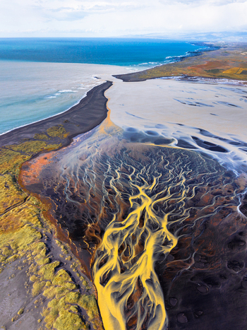 Aerial view of a glacial river delta with intricate braided channels carrying golden sediment across black volcanic sand plains meeting the Atlantic Ocean in Iceland. Aerial view of a glacial river delta with intricate braided channels carrying golden sediment across black volcanic sand plains meeting the Atlantic Ocean in Iceland.