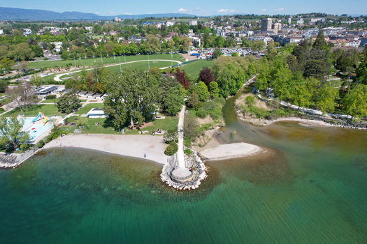 Aerial view of the scenic lakefront featuring a sandy beach lush green parkland and the river mouth flowing into the clear turquoise waters of Lake Geneva in Morges Vaud Switzerland. Aerial view of the scenic lakefront featuring a sandy beach lush green parkland and the river mouth flowing into the clear turquoise waters of Lake Geneva in Morges Vaud Switzerland.