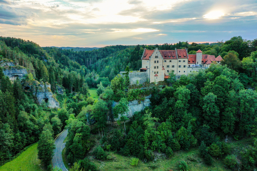 Aerial view of Burg Rabenstein perched on a limestone cliff surrounded by lush green forests and a winding road under a soft sunset sky Ahorntal Bayern Germany.