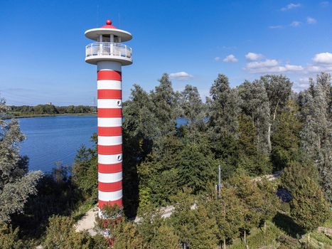 Aerial view of the striking red and white striped lighthouse standing tall against the lush greenery and calm waters Utopia Flevoland Netherlands. Aerial view of the striking red and white striped lighthouse standing tall against the lush greenery and calm waters Utopia Flevoland Netherlands.