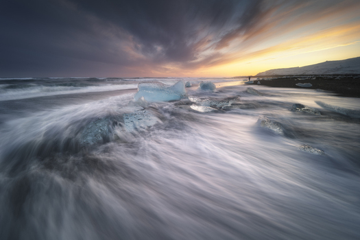 View of Diamond Beach featuring icebergs on black sand with long exposure waves under a dramatic sunset sky Sveitarfelagid Hornafjordur Iceland.