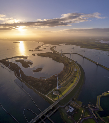 Aerial view of wind turbines situated along a coastal dike at sunset with calm water and golden light reflecting on the surface in the Netherlands. Aerial view of wind turbines situated along a coastal dike at sunset with calm water and golden light reflecting on the surface in the Netherlands.