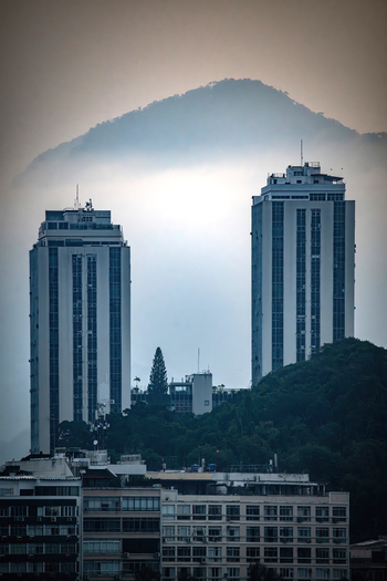 View of two modern skyscrapers rising above lush green hills with a misty mountain peak in the background Rio de Janeiro State of Rio de Janeiro Brazil. View of two modern skyscrapers rising above lush green hills with a misty mountain peak in the background Rio de Janeiro State of Rio de Janeiro Brazil.