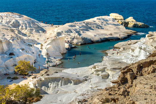 View of Sarakiniko Beach with its unique white volcanic rock formations turquoise water and people swimming and sunbathing under a clear sky in Milos Greece. View of Sarakiniko Beach with its unique white volcanic rock formations turquoise water and people swimming and sunbathing under a clear sky in Milos Greece.