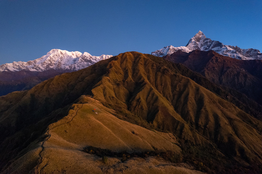 Aerial view of the majestic Machapuchare peak and the surrounding Annapurna mountain range at golden hour with grassy ridges and snow capped summits in Nepal.