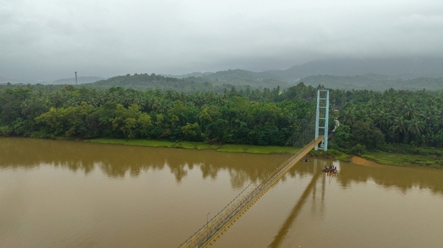Aerial view of the suspension bridge spanning the Sharavathi River surrounded by lush green palm trees and dense vegetation under an overcast sky in Honnavar Karnataka India.