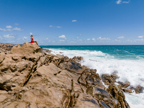 Aerial view of a woman in a red dress sitting on a rock at Injidup Natural Spa with turquoise waves crashing against the rugged coastline in Yallingup Western Australia Australia. Aerial view of a woman in a red dress sitting on a rock at Injidup Natural Spa with turquoise waves crashing against the rugged coastline in Yallingup Western Australia Australia.