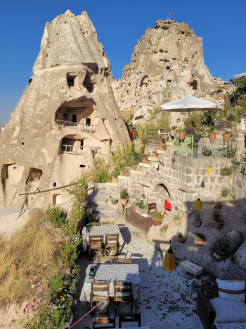 View of the ancient rock formations and cave dwellings of Cappadocia with a terrace cafe featuring tables and potted plants under a clear blue sky in Turkiye. View of the ancient rock formations and cave dwellings of Cappadocia with a terrace cafe featuring tables and potted plants under a clear blue sky in Turkiye.