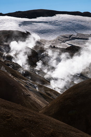View of geothermal steam rising from dark textured hills contrasting with a vast expanse of white glacier under a clear blue sky Hrunamannahreppur Iceland.