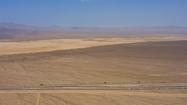 Aerial view of a long highway stretching through a vast arid desert landscape with distant mountains under a clear blue sky in the United States.