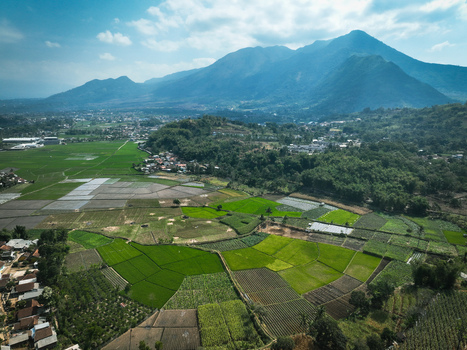 Aerial view of vibrant green rice fields and agricultural plots nestled in a valley beneath towering mountains in Garut West Java Indonesia. Aerial view of vibrant green rice fields and agricultural plots nestled in a valley beneath towering mountains in Garut West Java Indonesia.