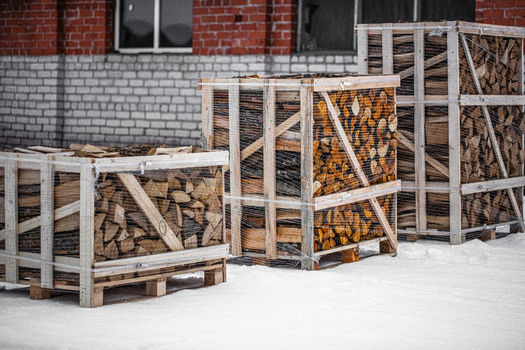 View of wooden crates filled with chopped firewood wrapped in plastic netting sitting on snow in front of a brick building in Latvia. View of wooden crates filled with chopped firewood wrapped in plastic netting sitting on snow in front of a brick building in Latvia.