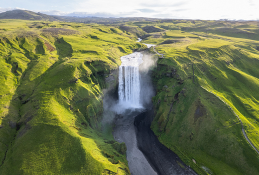 Aerial view of Skogafoss waterfall cascading down lush green cliffs into a river valley under soft sunlight Rangarthing eystra Iceland.