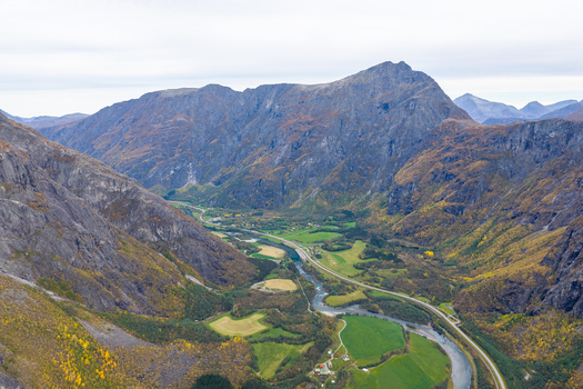 Aerial view of the Romsdalen valley featuring a winding river lush green fields and a road flanked by towering rugged mountains with autumn foliage in Andalsnes More og Romsdal Norway. Aerial view of the Romsdalen valley featuring a winding river lush green fields and a road flanked by towering rugged mountains with autumn foliage in Andalsnes More og Romsdal Norway.