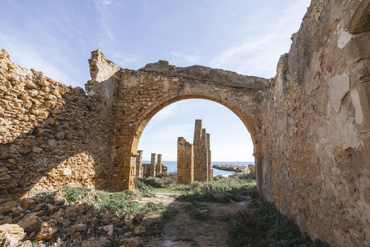 View of the historic stone ruins of Tonnara di Avola featuring a large archway and pillars overlooking the sea under a clear blue sky in Avola Sicily Italy. View of the historic stone ruins of Tonnara di Avola featuring a large archway and pillars overlooking the sea under a clear blue sky in Avola Sicily Italy.