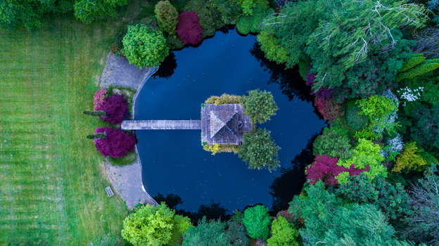 Aerial view of a circular pond featuring a wooden pavilion and bridge surrounded by vibrant green and purple foliage and a manicured lawn in Portland Oregon United States. Aerial view of a circular pond featuring a wooden pavilion and bridge surrounded by vibrant green and purple foliage and a manicured lawn in Portland Oregon United States.