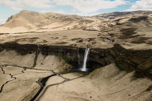Aerial view of Seljalandsfoss waterfall cascading over rocky cliffs into a pool surrounded by expansive dry golden plains and winding walking paths in Rangarthing eystra Iceland. Aerial view of Seljalandsfoss waterfall cascading over rocky cliffs into a pool surrounded by expansive dry golden plains and winding walking paths in Rangarthing eystra Iceland.