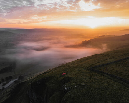 Aerial view of Winnats Pass at sunrise with a red tent on the grassy hillside above a valley filled with morning mist in Hope Valley England United Kingdom. Aerial view of Winnats Pass at sunrise with a red tent on the grassy hillside above a valley filled with morning mist in Hope Valley England United Kingdom.
