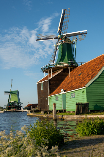 View of the historic Zaanse Schans windmills with green wooden structures and orange tiled roofs along the water under a clear blue sky in Zaandam Noord Holland Netherlands.