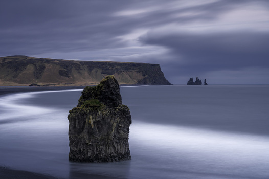 View of the Arnardrangur sea stack on a black sand beach with Reynisdrangar rock formations in the distance under a moody sky Arnardrangur Myrdalshreppur Iceland.