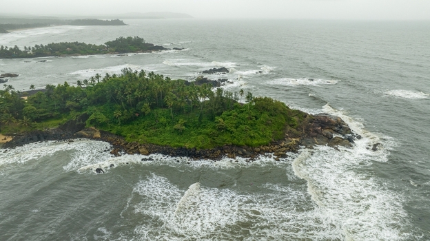 Aerial view of Palolem Beach featuring a lush green rocky headland surrounded by crashing waves and the vast Arabian Sea in Canacona Goa India.