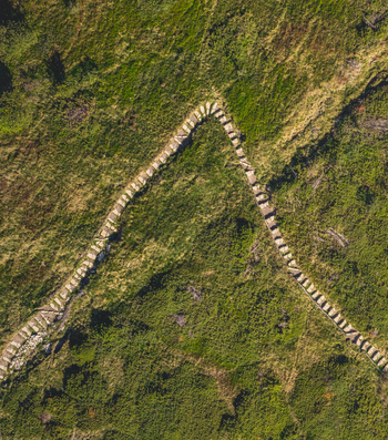 Aerial view of a sharp hairpin bend on a stone hiking trail crossing a green mountain slope covered with grass and dense shrubs in France.