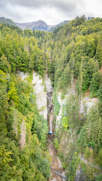 Aerial view of Grandvillard Waterfall cascading down a steep rocky cliff surrounded by dense green forest and alpine mountains under a cloudy sky in Grandvillard Fribourg Switzerland.