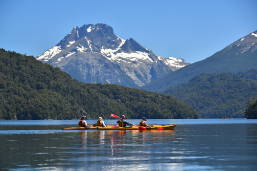 View of people kayaking on the calm waters of Nahuel Huapi Lake with snow capped mountains and lush forests in San Carlos de Bariloche Rio Negro Argentina. View of people kayaking on the calm waters of Nahuel Huapi Lake with snow capped mountains and lush forests in San Carlos de Bariloche Rio Negro Argentina.