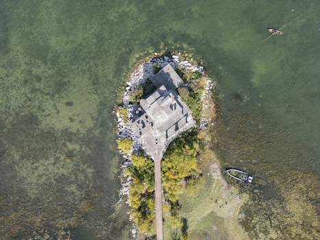 Aerial view of a building on a small peninsula extending into Lake Shkodra surrounded by clear green water with a wooden walkway and a small boat nearby in Shiroka Shkoder County Albania. Aerial view of a building on a small peninsula extending into Lake Shkodra surrounded by clear green water with a wooden walkway and a small boat nearby in Shiroka Shkoder County Albania.
