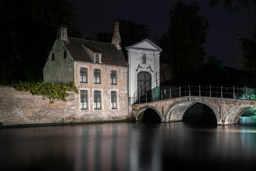 View of the historic Sas Huis and a stone arch bridge reflecting in the calm canal waters at night in Bruges Flanders Belgium.