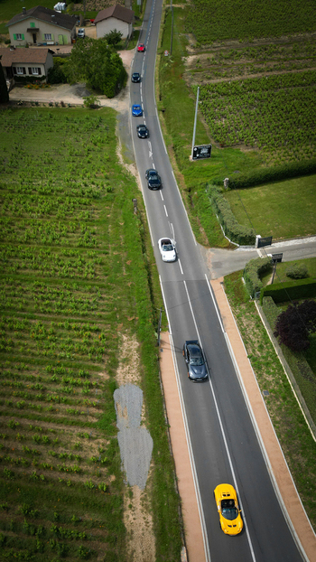 Aerial view of sports cars driving on a rural road through lush green vineyards and houses in La Chapelle de Guinchay Bourgogne Franche Comte France.
