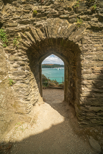 View of the Fowey River framed by a historic stone archway with boats sailing on the water and the town of Fowey in the distance in Fowey Cornwall United Kingdom. View of the Fowey River framed by a historic stone archway with boats sailing on the water and the town of Fowey in the distance in Fowey Cornwall United Kingdom.