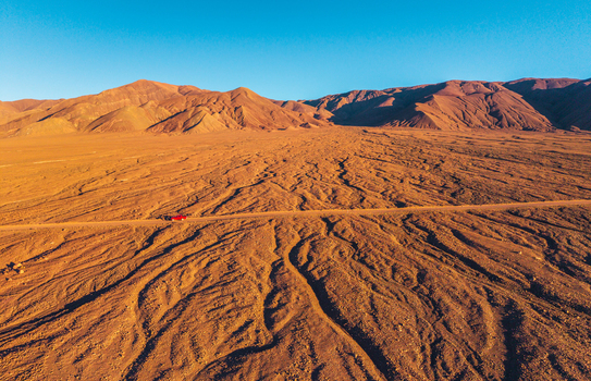 Aerial view of a red car driving along a dusty road through the arid Atacama Desert with rugged mountains under a clear blue sky in San Pedro de Atacama Antofagasta Chile. Aerial view of a red car driving along a dusty road through the arid Atacama Desert with rugged mountains under a clear blue sky in San Pedro de Atacama Antofagasta Chile.